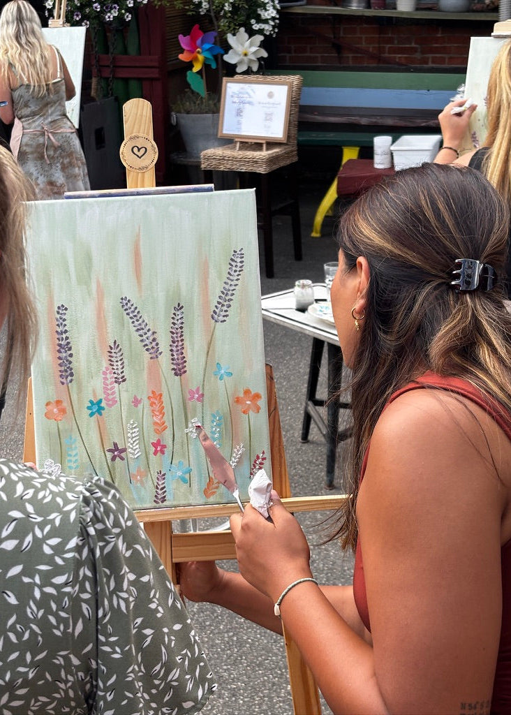 A woman painting a floral art piece on an easel at an outdoor craft or market event, with other people and tables visible in the background.