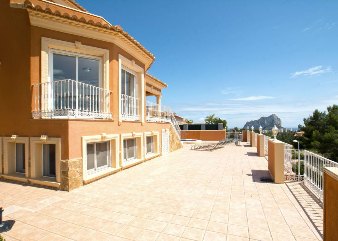 A large tiled terrace with a modern orange stucco building featuring white railings, set against a clear blue sky with distant mountains.