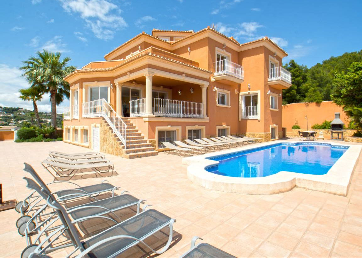 A large orange stucco villa with multiple balconies, a tiled outdoor patio, rows of lounge chairs, and a bright blue swimming pool under a blue sky with clouds.