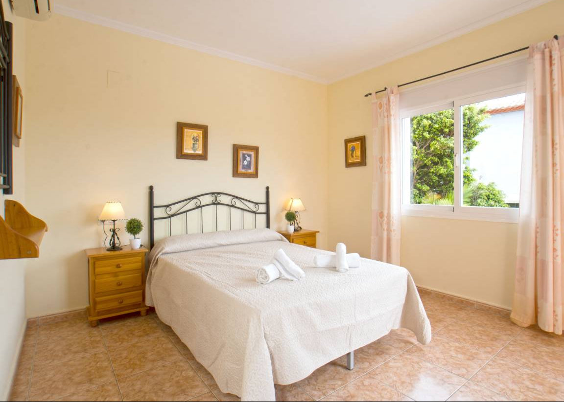 A double bed with white bedding and a black metal headboard in a bedroom with pale yellow walls, wooden nightstands, lamps, and a window with pink curtains.
