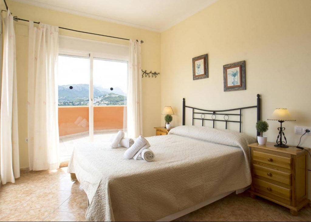 A cozy bedroom with a neatly made bed featuring beige bedding, a black metal headboard, and a wooden nightstand beside it. The room has pale yellow walls, white curtains, and a window looking out onto a balcony with a view of hills.
