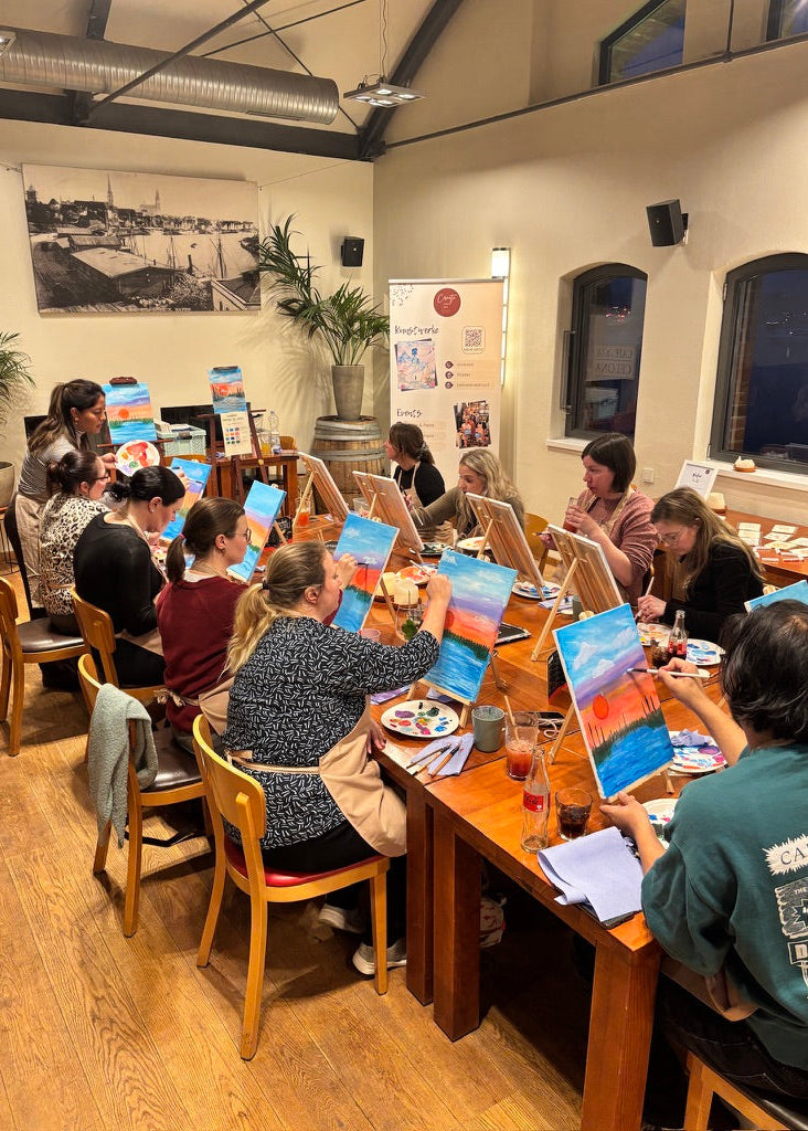 A group of people seated around a long wooden table in a warmly lit indoor studio or workshop, each holding a small landscape painting with blue sky and colorful sunset tones. The room has wood floors, cream walls, windows, and framed wall art.