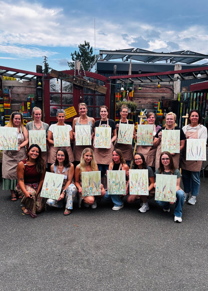 A group of women wearing matching tan aprons pose outdoors in front of a greenhouse, each holding up a small painted artwork. The scene includes a paved ground, greenhouse structure, and cloudy sky.