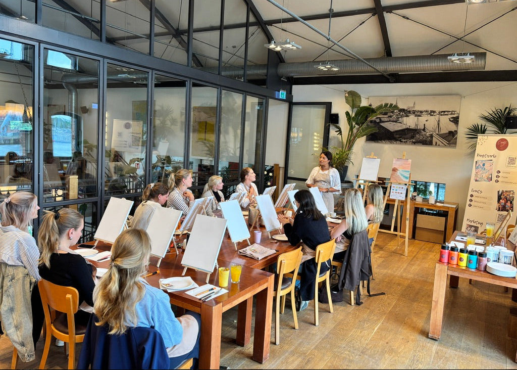 A group of people seated at long wooden tables in a bright studio or classroom, each holding a large white sketchbook or canvas, with an instructor standing near easels and a paint supply table in the background.