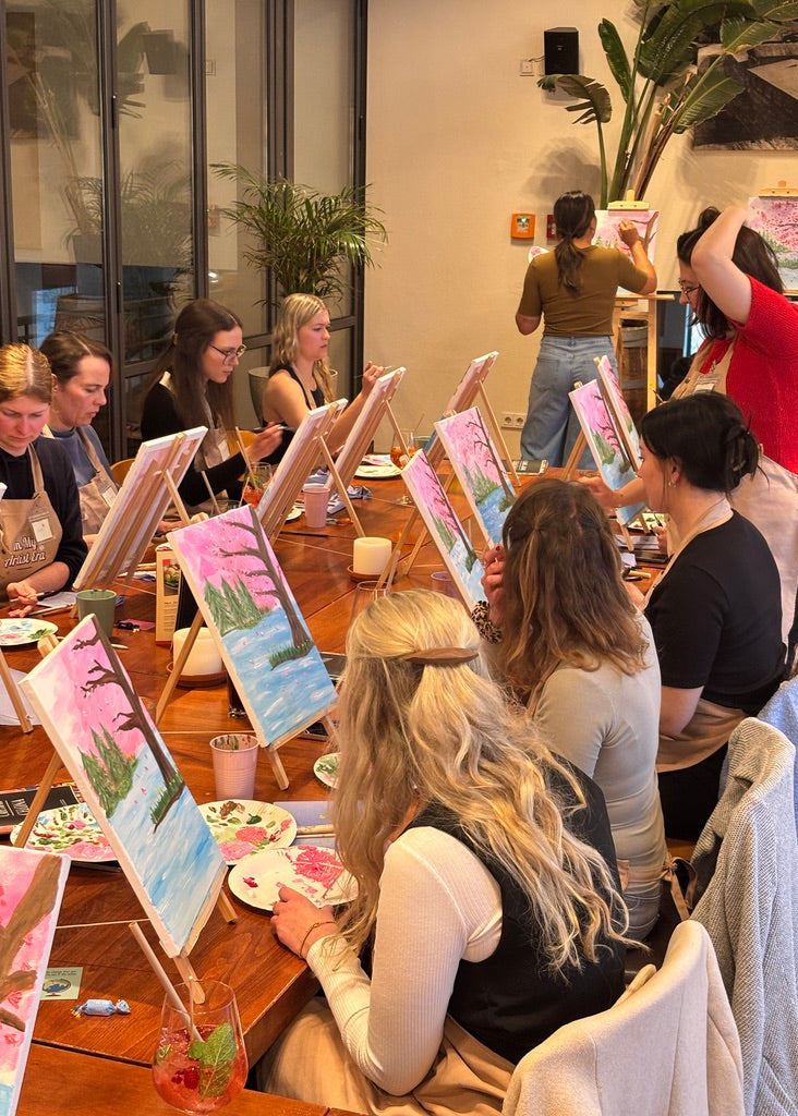 A group of people seated around a wooden table in a studio, each with a small landscape painting on an easel in front of them. Several people are standing and painting in the background, with large windows, glass doors, and indoor plants visible.