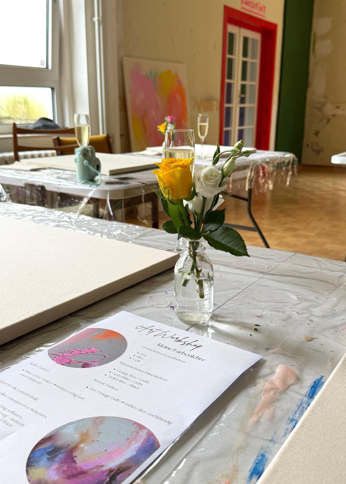 A small clear glass vase holding yellow and white roses sits on a table covered with a clear plastic sheet in a bright indoor room. A printed sheet with colorful circular artwork and handwritten text lies in front, with other tables, chairs, and artwork visible in the background.