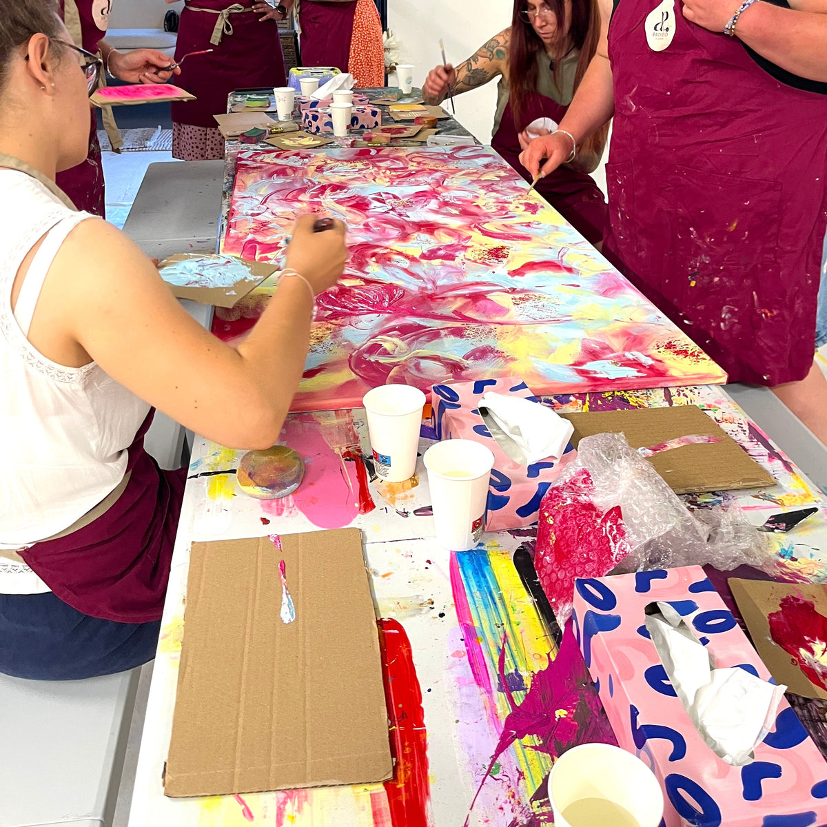 A group of people painting on a large canvas table during an art workshop or class. The table is covered with paint, cups, brushes, and cardboard boxes, and the participants wear maroon aprons.