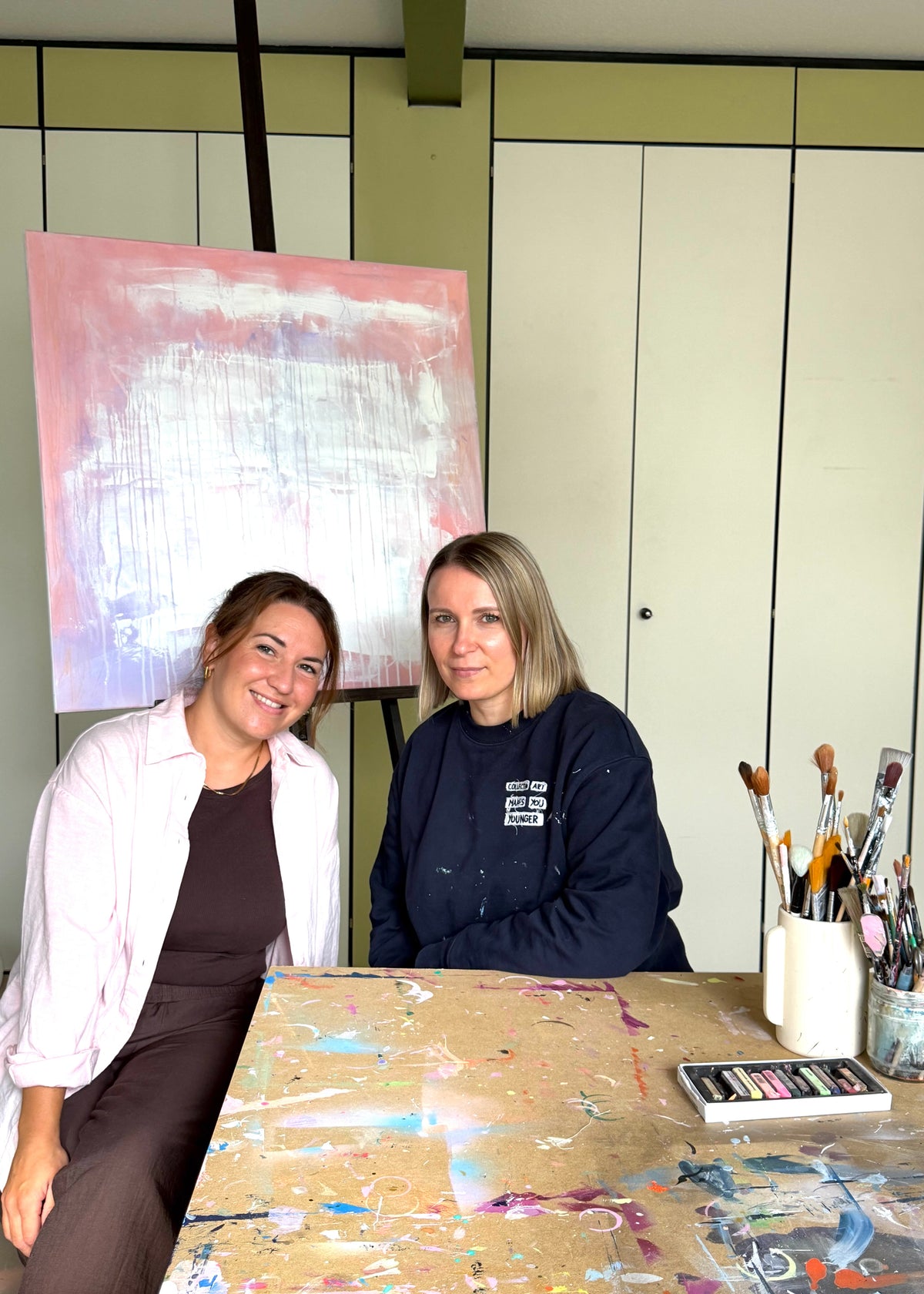 Two women sitting at a paint-splattered table in an art studio, with a large abstract pink and white painting on an easel behind them and paintbrushes in containers on the table.