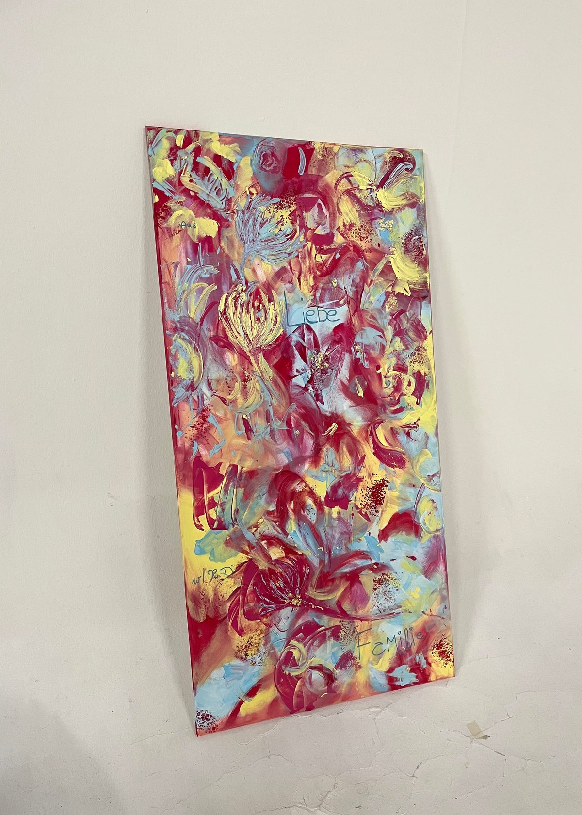 An abstract vertical canvas painting with a vibrant mix of red, yellow, blue, and pink brushstrokes, leaning against a plain off-white wall.