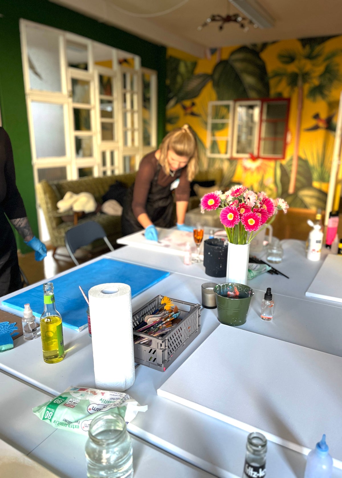 A lifestyle scene in a creative studio or classroom with a tropical mural on the wall. A woman is seen working at a table with art supplies, including a roll of white paper, a vase of pink flowers, and blue sheets on the table.