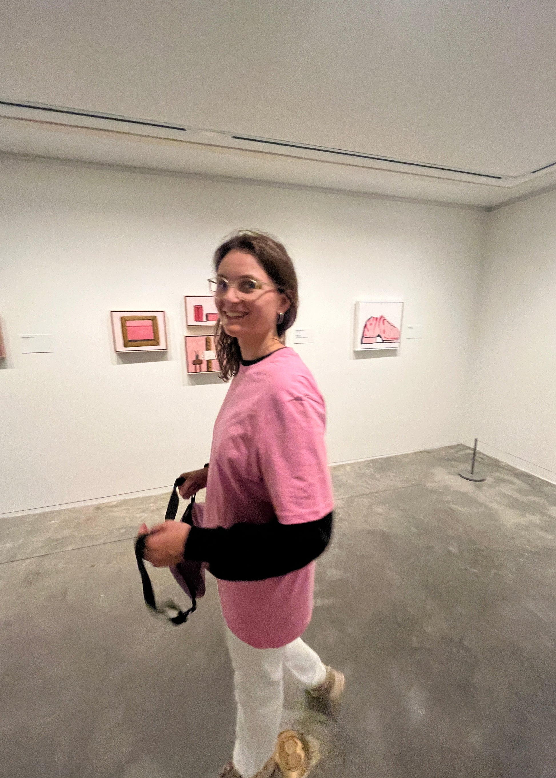 Person wearing a pink jacket and white pants standing in a room with gray walls and a door.