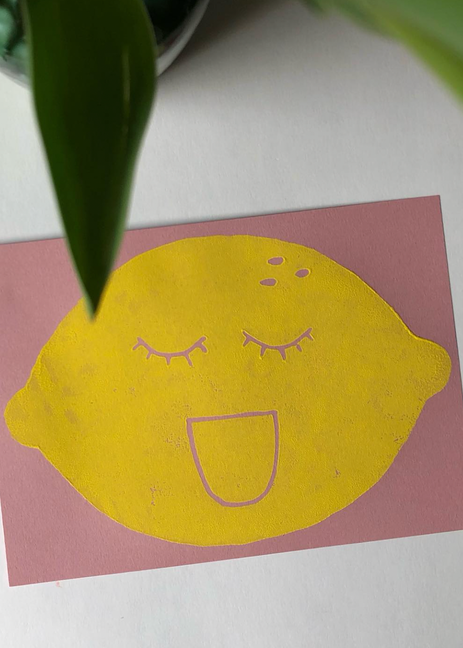 Yellow lemon with a face on a pink card on a white surface with green leaves.