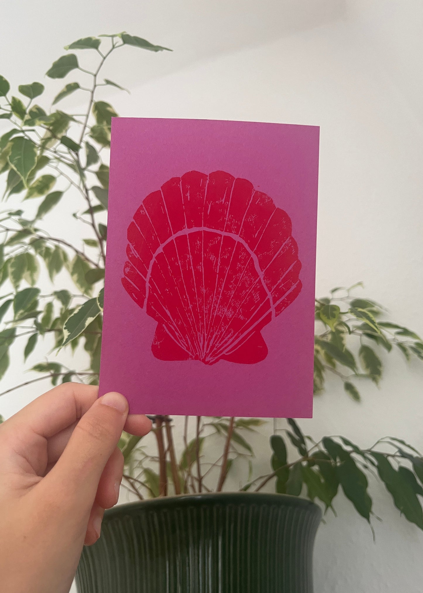 Hand holding a pink card with a red shell design against a white background with green leaves.