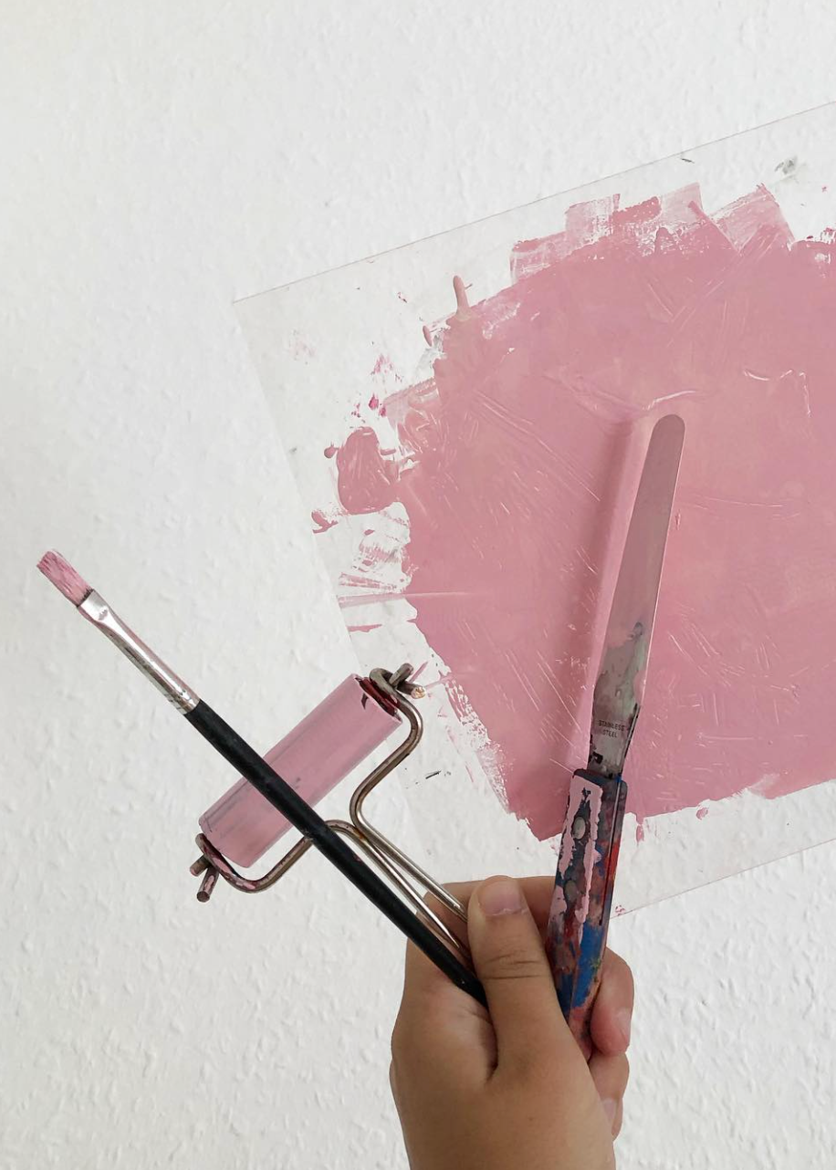 Hand holding a paintbrush and roller over a pink paint swatch on a white background