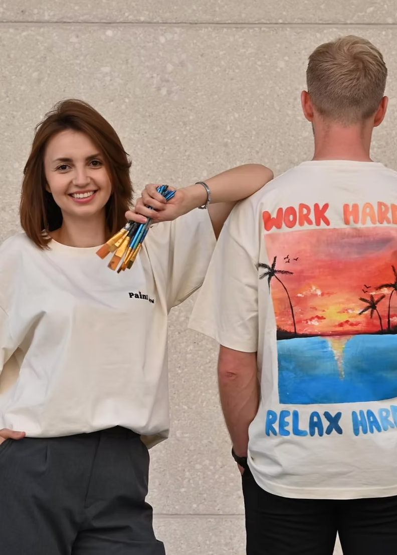 Two people wearing white t-shirts with artistic designs, one with paintbrushes and the other with a sunset design.