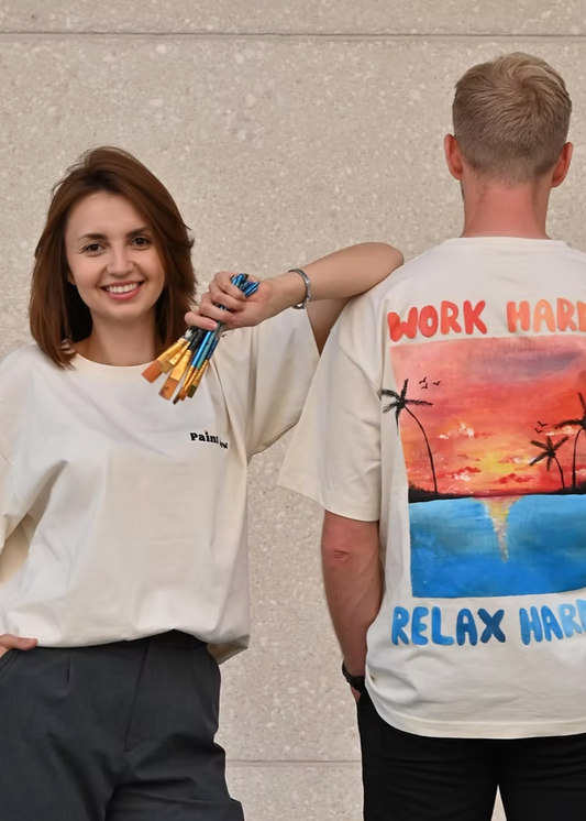 Two people wearing white t-shirts with artistic designs, one with paintbrushes and the other with a sunset design.