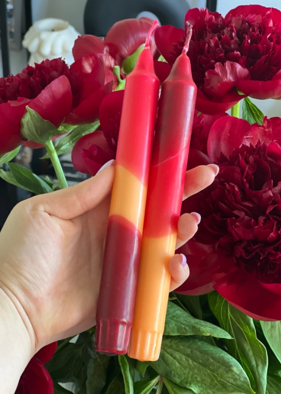 Hand holding two red and orange gradient candles in front of red flowers