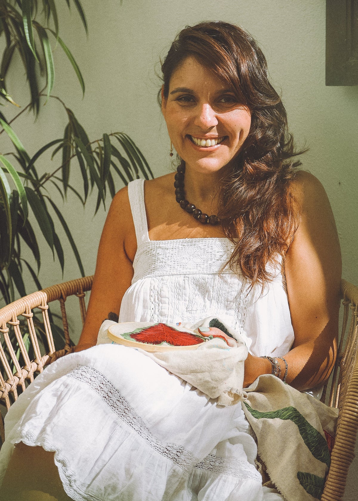 A smiling woman with long brown hair sits in a wicker chair, wearing a white sleeveless dress and holding a white blanket with a red and green pattern.