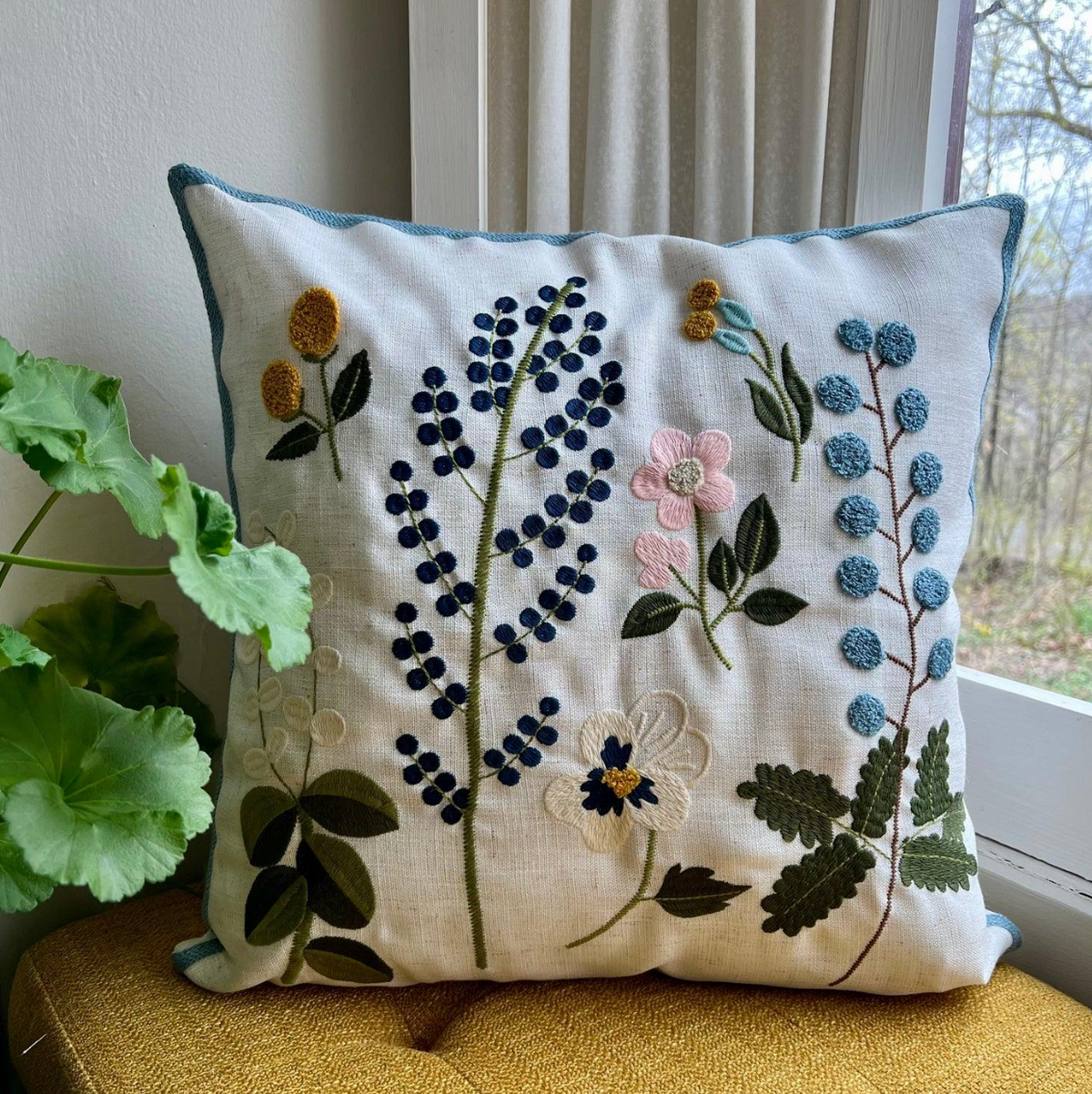 A square decorative throw pillow with embroidered wildflowers on a cream linen background, styled on a yellow cushion next to a window and green plant leaves.
