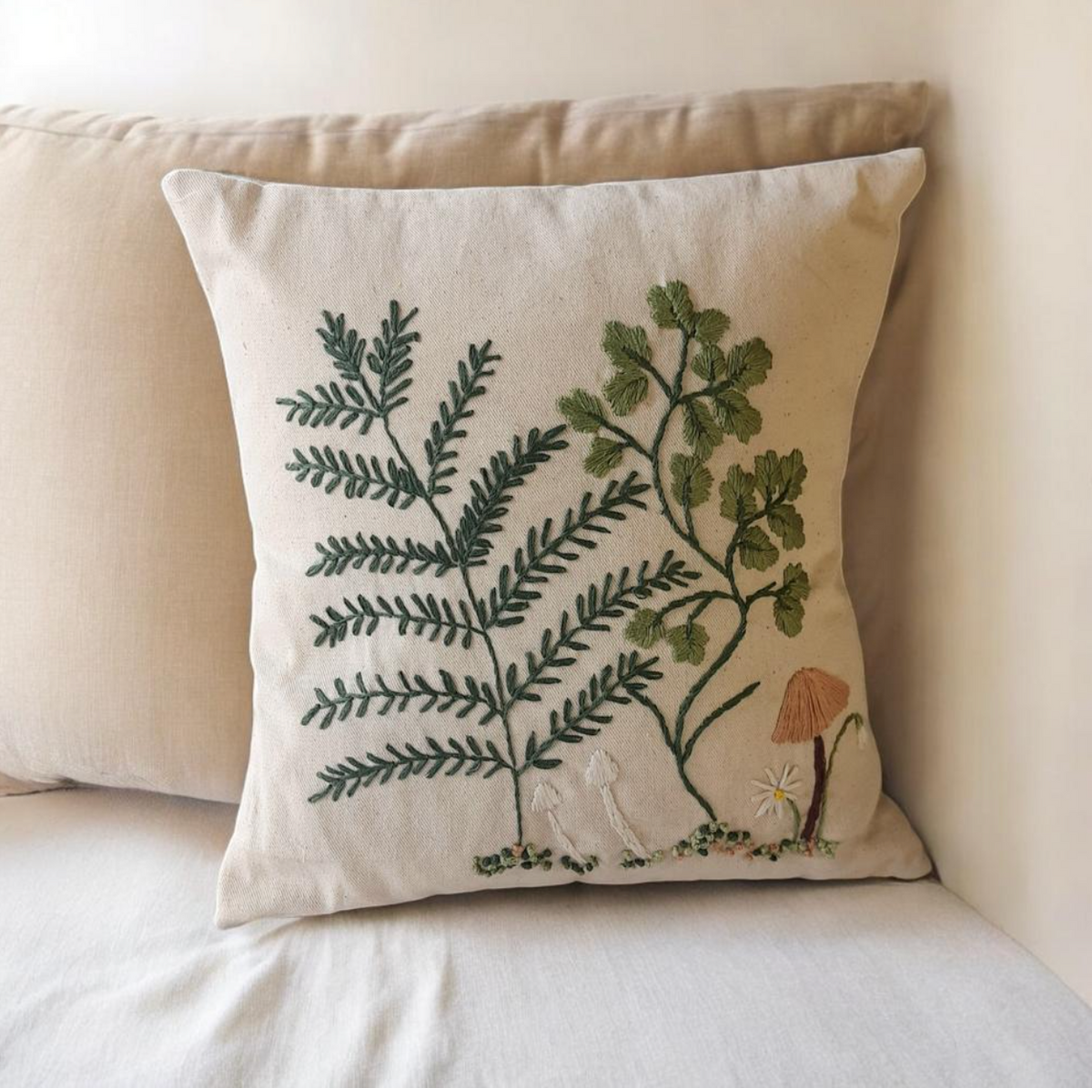 A square beige throw pillow with detailed botanical embroidery of green ferns, a small mushroom, and a white daisy, styled on a bed with a neutral cream background.