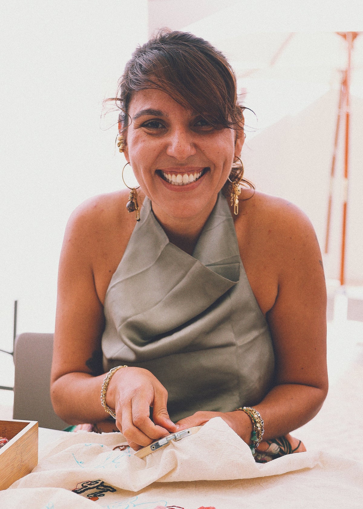 A smiling woman with dark hair pulled back sits at a table, wearing a draped olive green halter top. She is holding a white fabric with colorful embroidered lettering, with a wooden box and more fabric visible in the foreground.