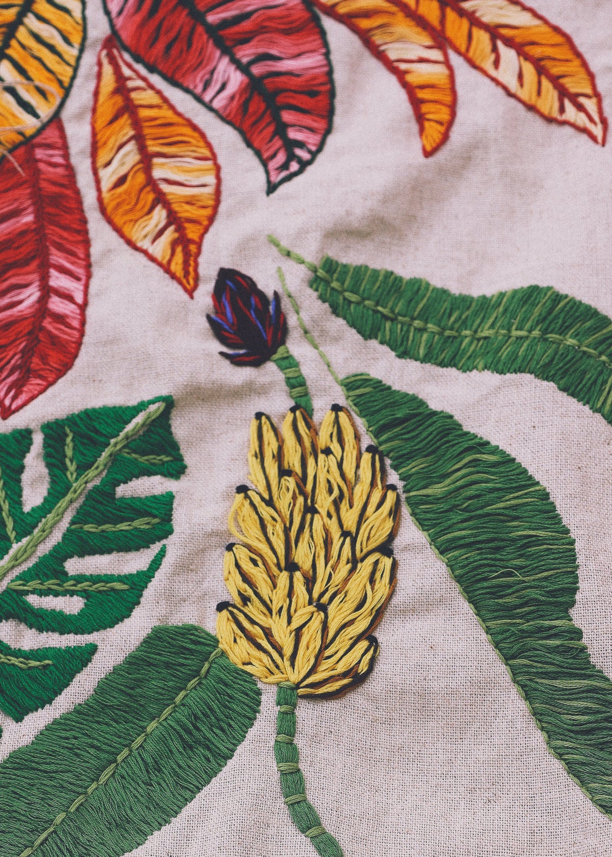 Close-up of a woven textile featuring colorful embroidered tropical leaves and a yellow flower on a light beige background.