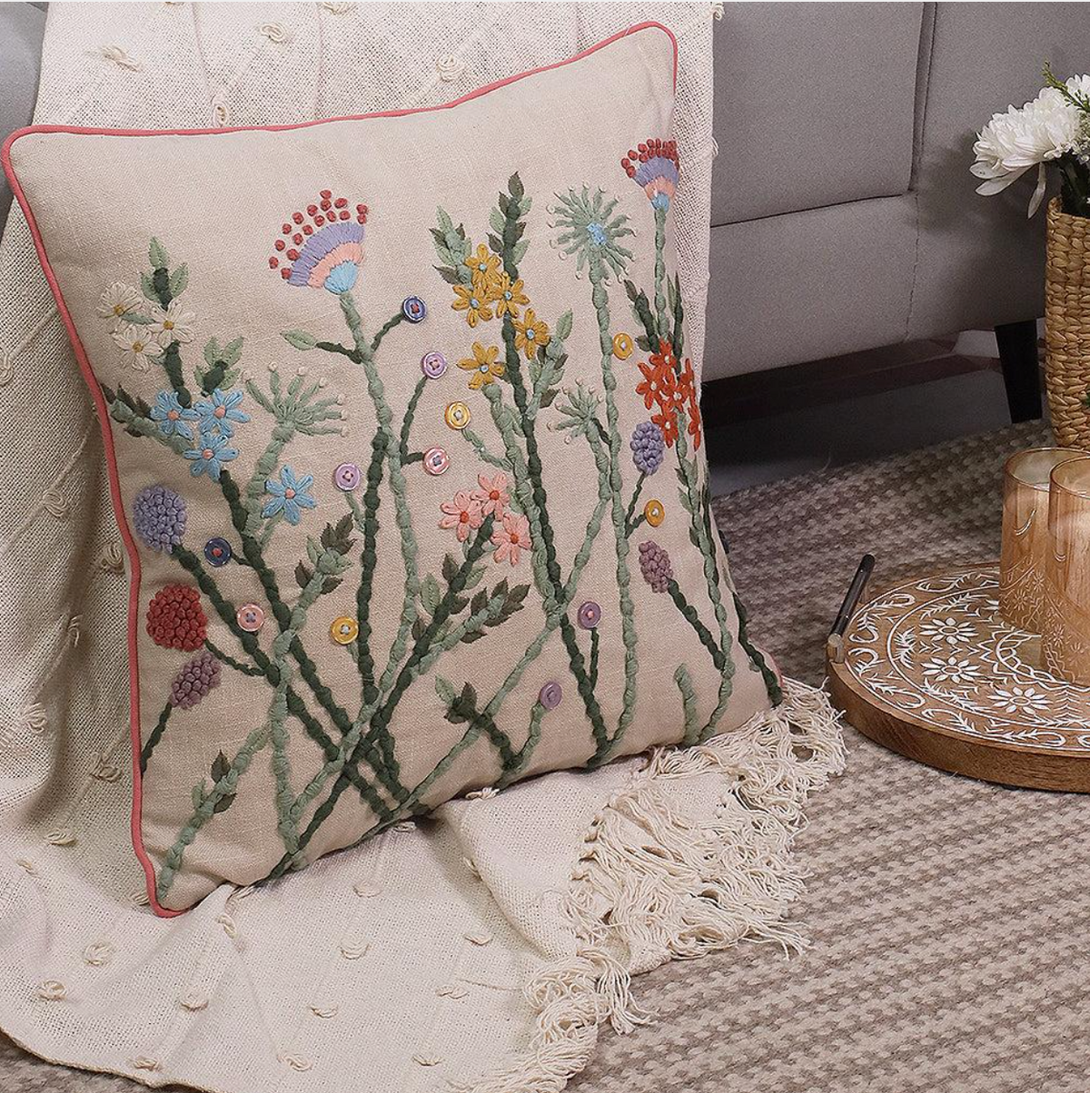 A square decorative throw pillow with a beige linen-like base, featuring colorful embroidered wildflowers and green stems, finished with a thin red piping border. The pillow is styled on a cream textured throw blanket in a cozy living room setting.