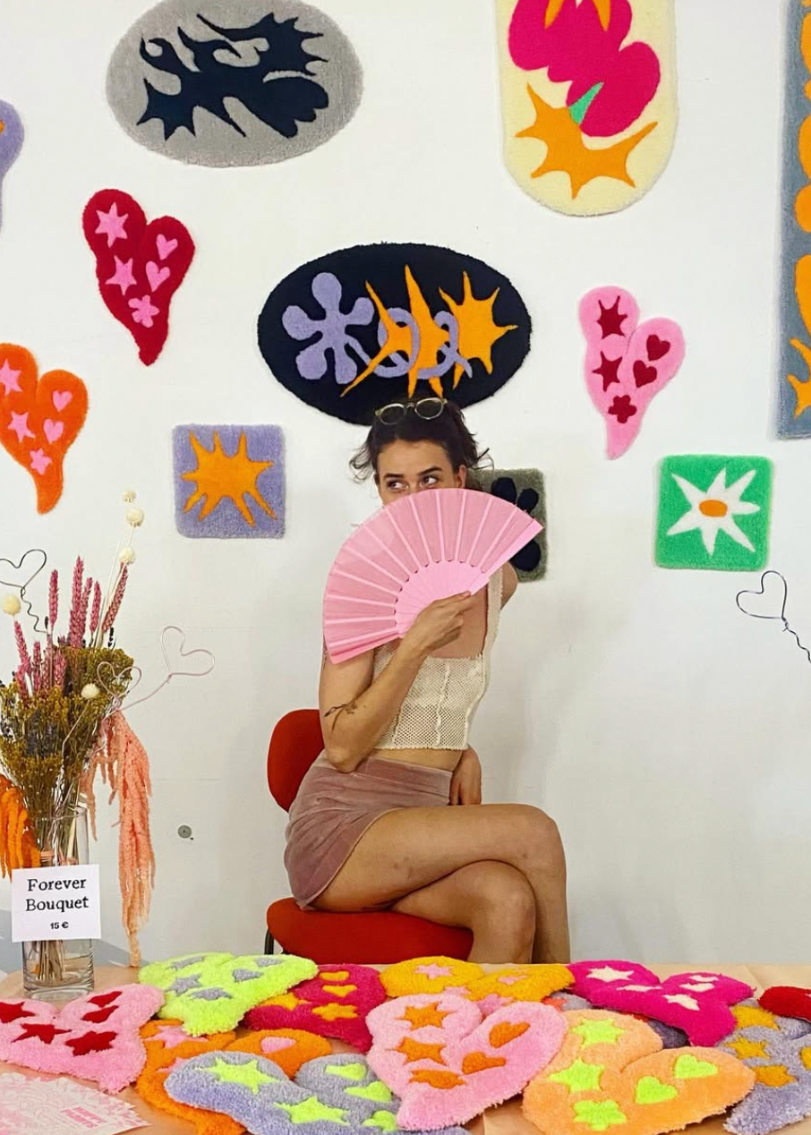 Woman holding a pink fan in front of her face with colorful abstract art on the wall behind her.