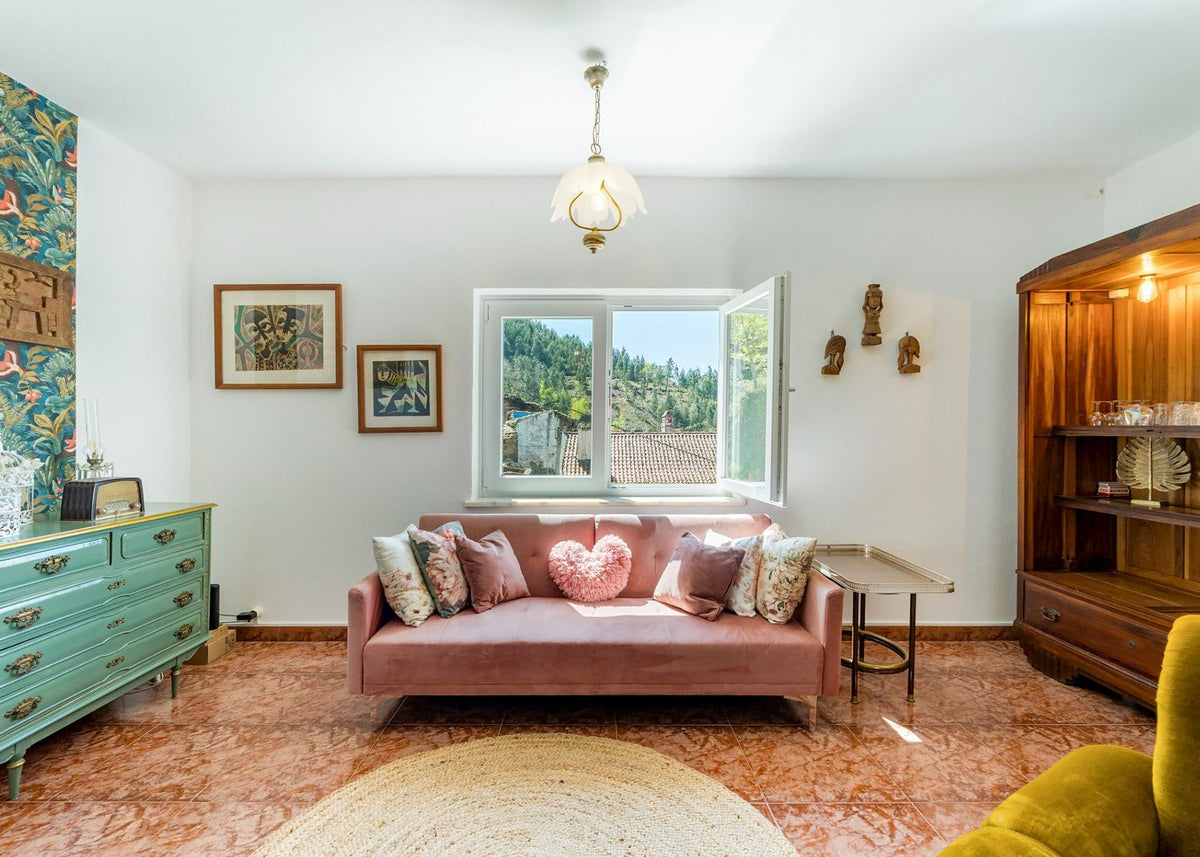 A styled living room featuring a dusty rose velvet sofa with patterned throw pillows, including a heart-shaped pillow, set against a white wall with framed art. The room has terracotta tiled floors, a turquoise wooden dresser on the left, and a large wooden hutch on the right.