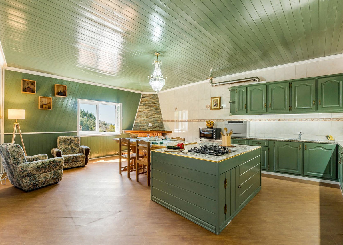 A spacious kitchen with sage green cabinets and matching green island, featuring a wood-look floor, white tiled backsplash, and a central stone chimney stack.
