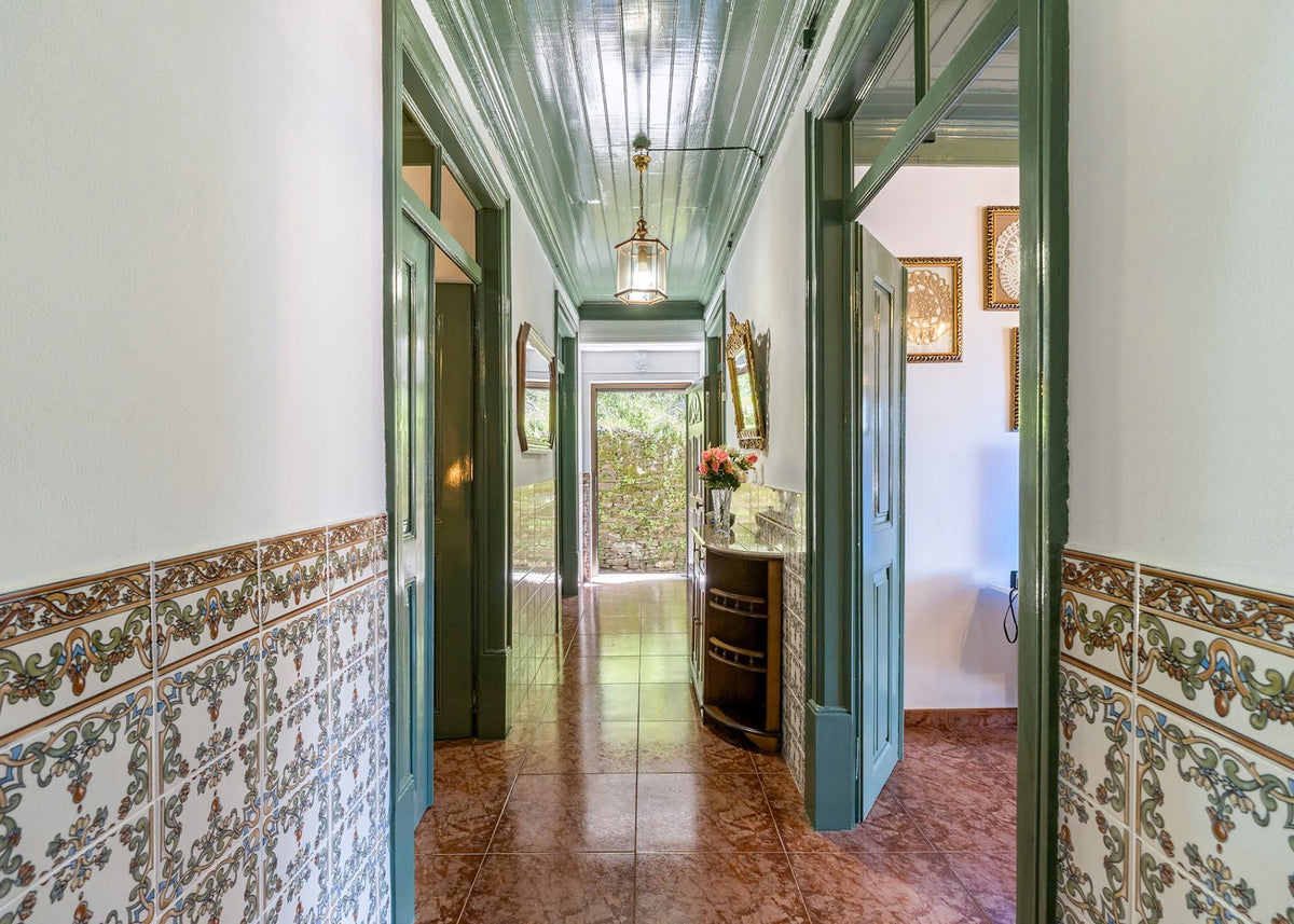 A long hallway with decorative patterned tile wainscoting, dark green trim, a corrugated metal ceiling, and a hanging lantern light. The floor is reddish-brown tile, and an open doorway on the right reveals another room with framed wall art.