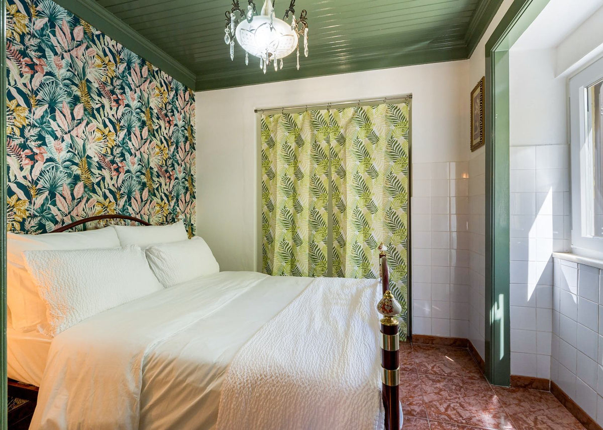 A styled bedroom interior featuring a bed dressed in white bedding, a green patterned wallpaper accent wall, matching green and white curtains, white tiled walls, and a window on the right side.