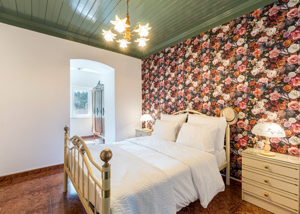 A bedroom interior featuring a bed dressed in white bedding, a floral wallpaper accent wall, a light wood nightstand with a lamp, and a green paneled ceiling.