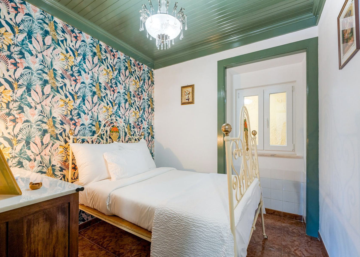 A styled bedroom interior featuring a bed with white bedding and a gold metal frame, set against a wall with colorful tropical floral wallpaper. The room has green trim, a green paneled ceiling with a hanging crystal chandelier, and a wooden nightstand in the foreground.