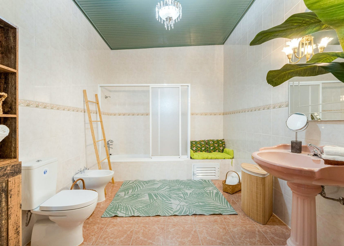 A tropical-themed bathroom featuring a pink pedestal sink, a green palm-patterned rug, a green and yellow cushion on a white bathtub, and a light green ceiling.