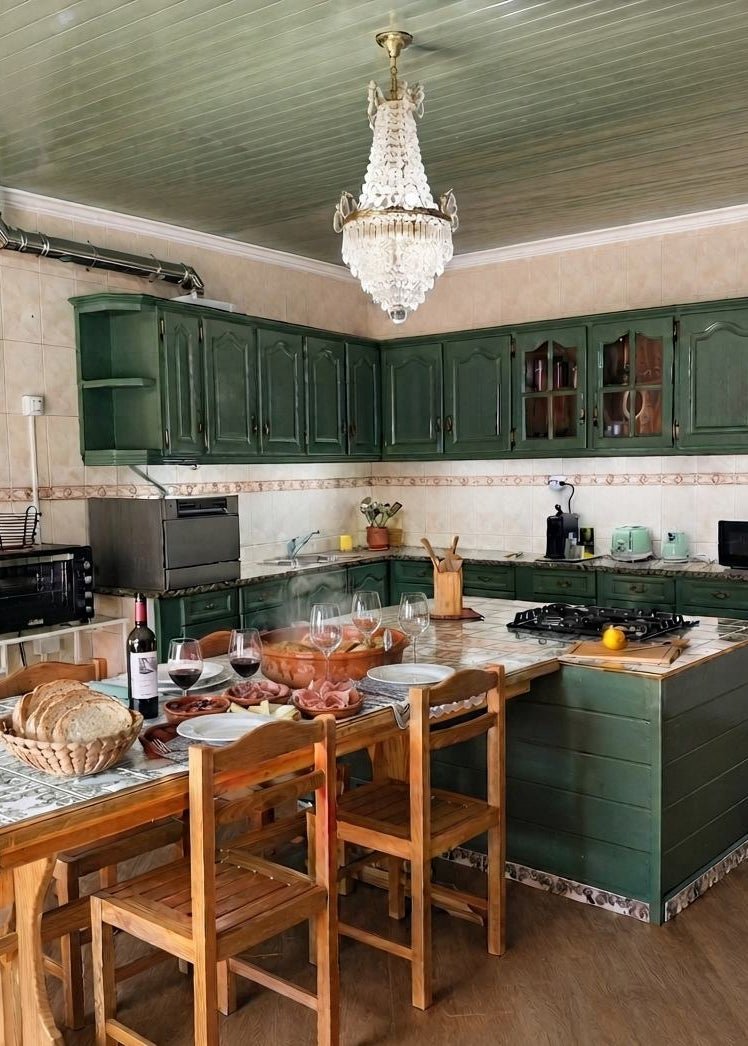 A rustic kitchen interior with dark green wooden cabinets and matching lower kitchen units, a wooden dining table with chairs, a crystal chandelier, tiled walls, and a countertop with food and dining items.
