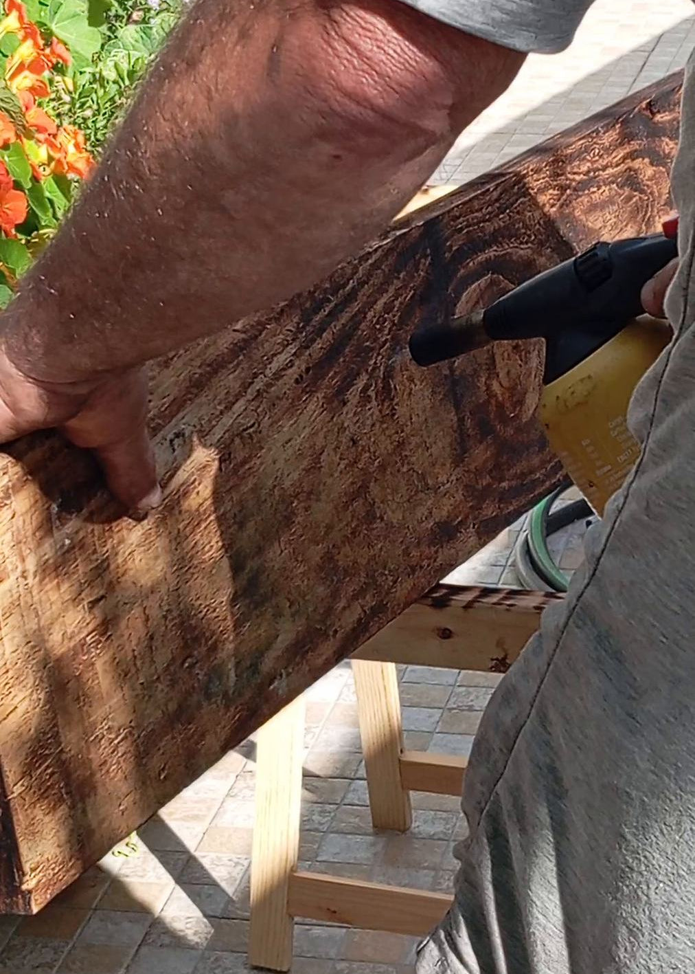 A person outdoors using a black heat gun to apply heat to a large, dark brown wood board resting on a wooden stand.