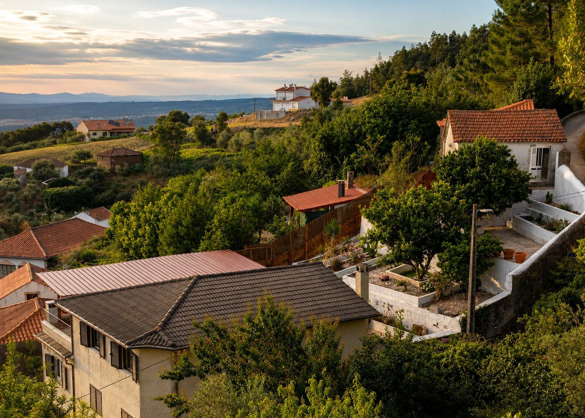 A scenic view of a hillside village with white houses, terracotta and dark tiled roofs, dense green trees, and distant mountains under a soft sunset sky.