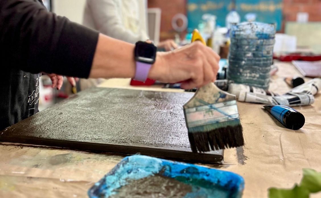 Person painting a canvas with a brush on a table with art supplies.