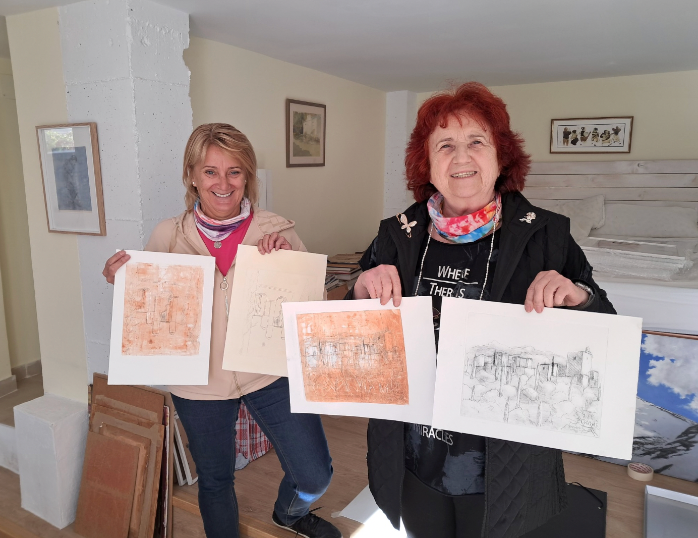 Two women holding architectural drawings in a room with framed pictures on the wall.