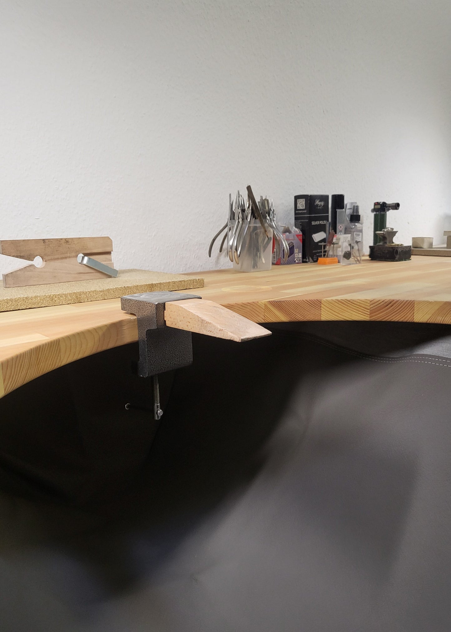 A curved wooden workbench with a black leather or vinyl seat, shown in a workshop setting with tools in the background.