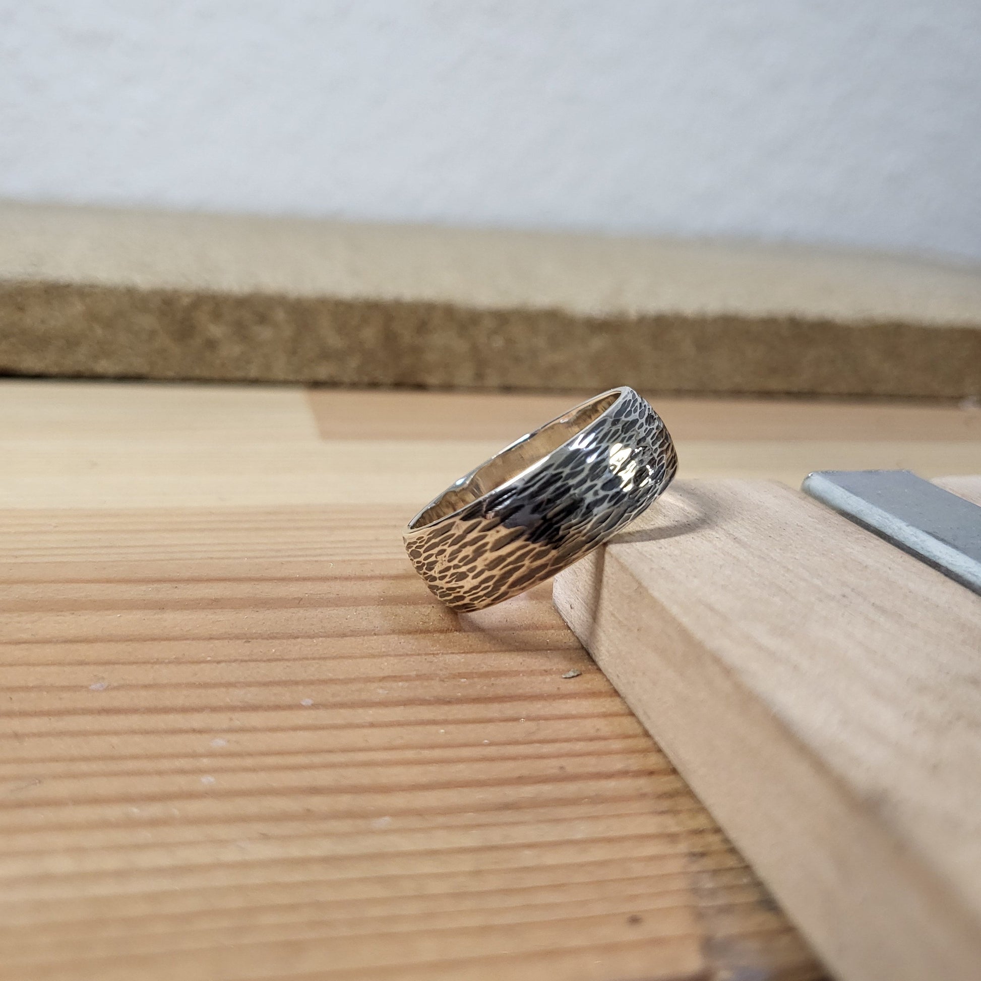 A textured silver ring with a hammered or cross-hatched pattern, resting on a light-colored wooden surface.