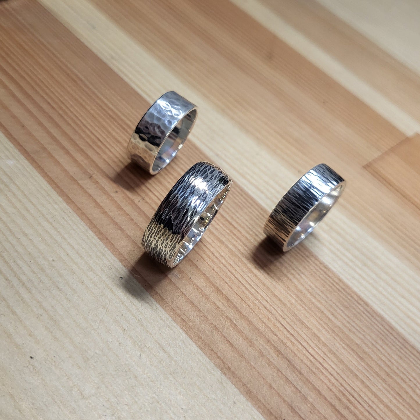 Three silver rings with textured finishes displayed on a light wood grain surface.