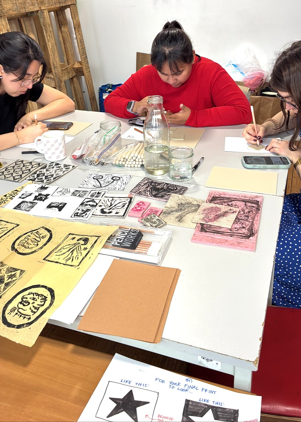 Three people seated around a table working on art prints and stamps in a studio setting. The table is covered with paper, ink blocks, brushes, and art supplies.