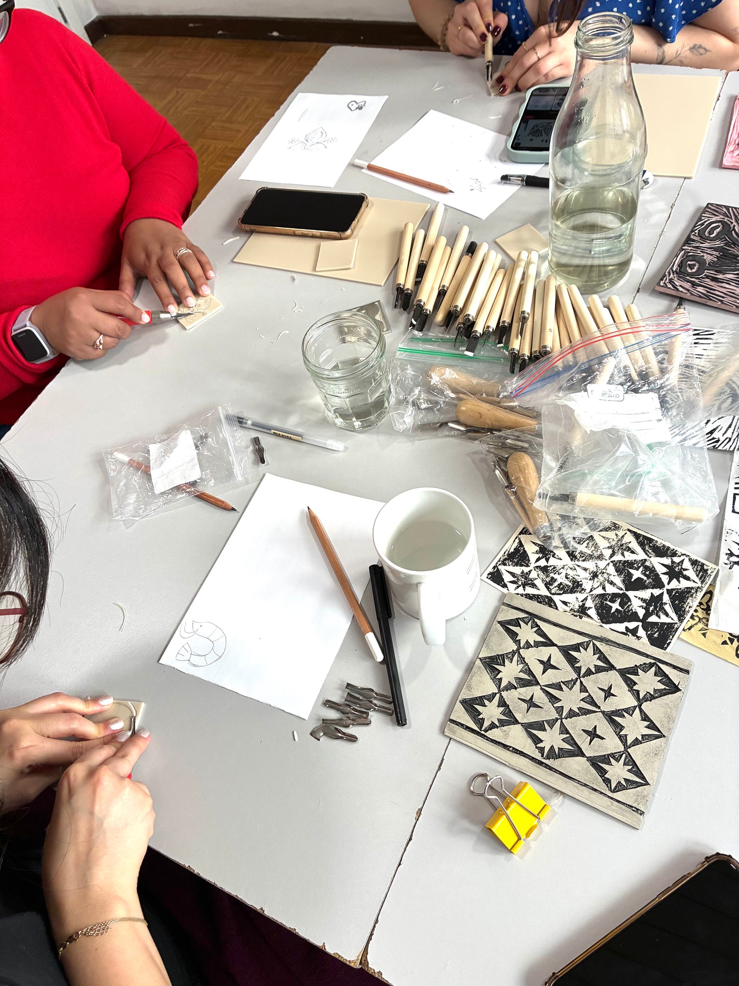 A tabletop scene showing multiple people working at a table with craft supplies, including patterned tiles, pencils, small tools, a glass bottle of water, and papers. The environment is a real indoor workspace with visible people's hands and partial bodies, so this is Lifestyle rather than Product/Packaging.