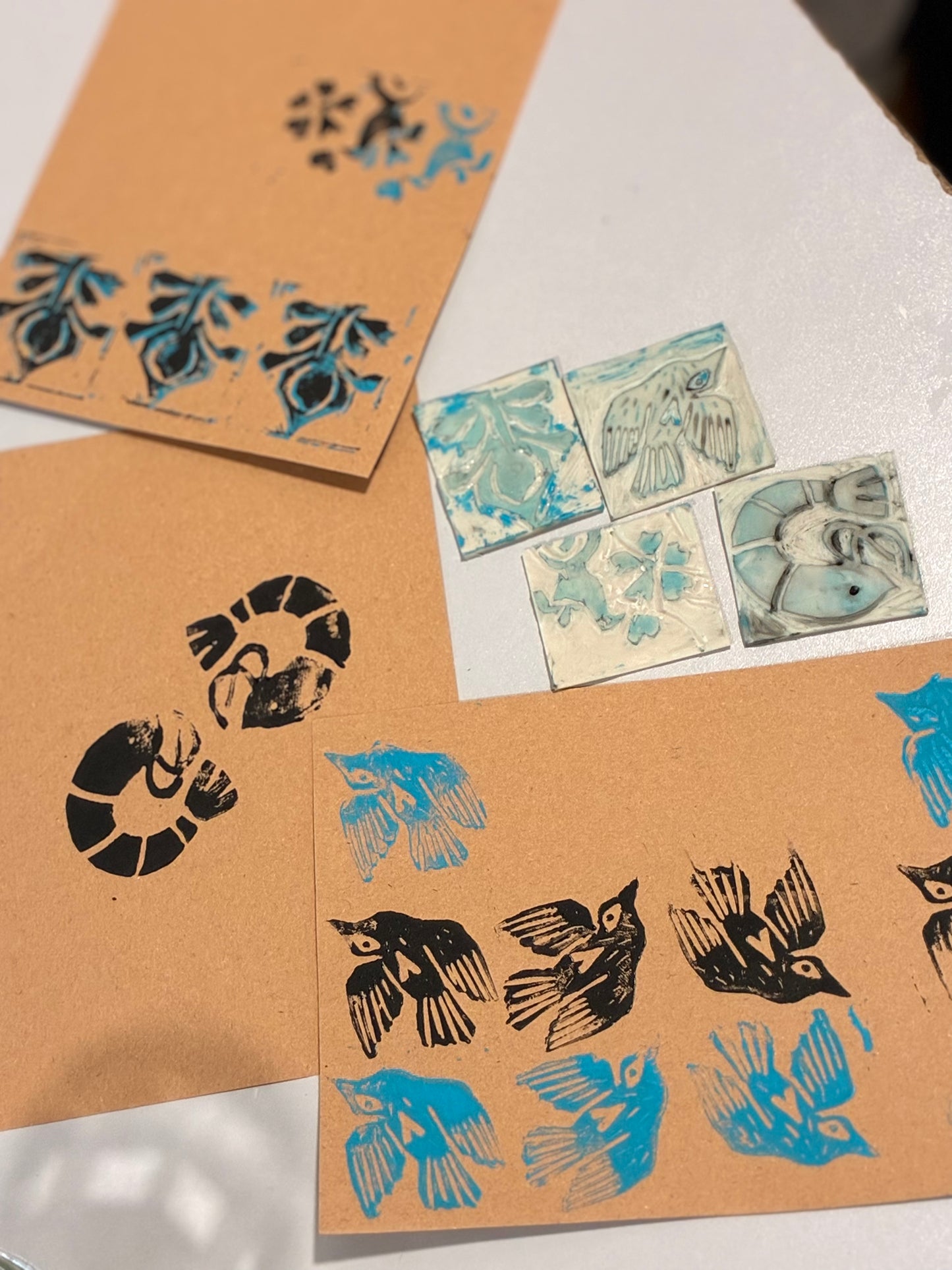 A flat-lay view of handmade block print cards and small ceramic tiles featuring blue and black bird and floral designs on a white surface.