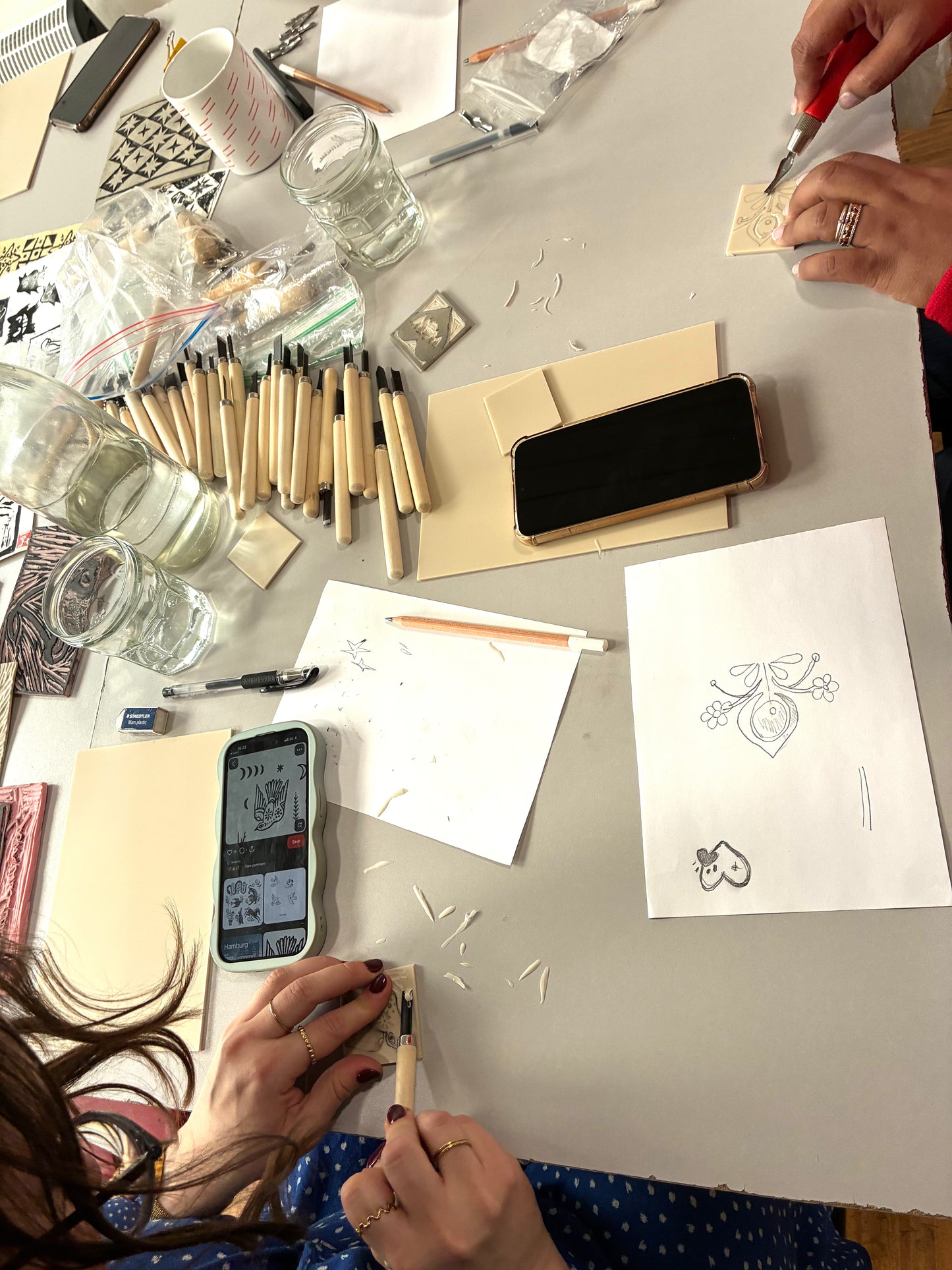 A top-down view of a craft table with multiple hands working on jewelry-making projects. The scene includes wooden carving tools, small brushes, glass jars, sketch papers with designs, and a smartphone showing a design app.