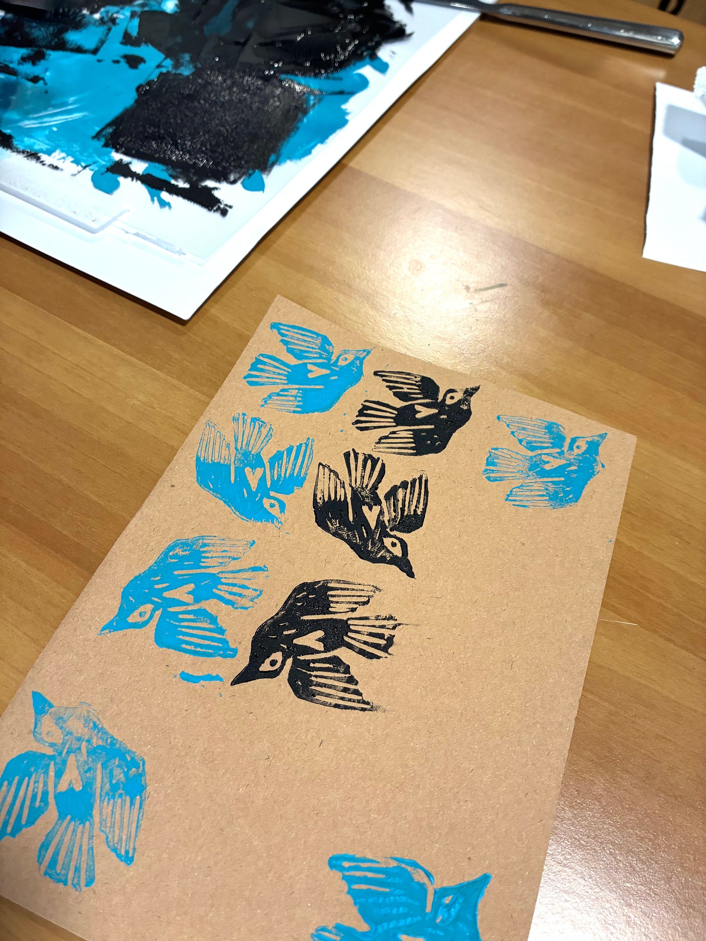 A sheet of kraft paper printed with repeated bird silhouettes in black and bright blue, photographed on a wooden tabletop. A paint palette and paper are partially visible in the background.