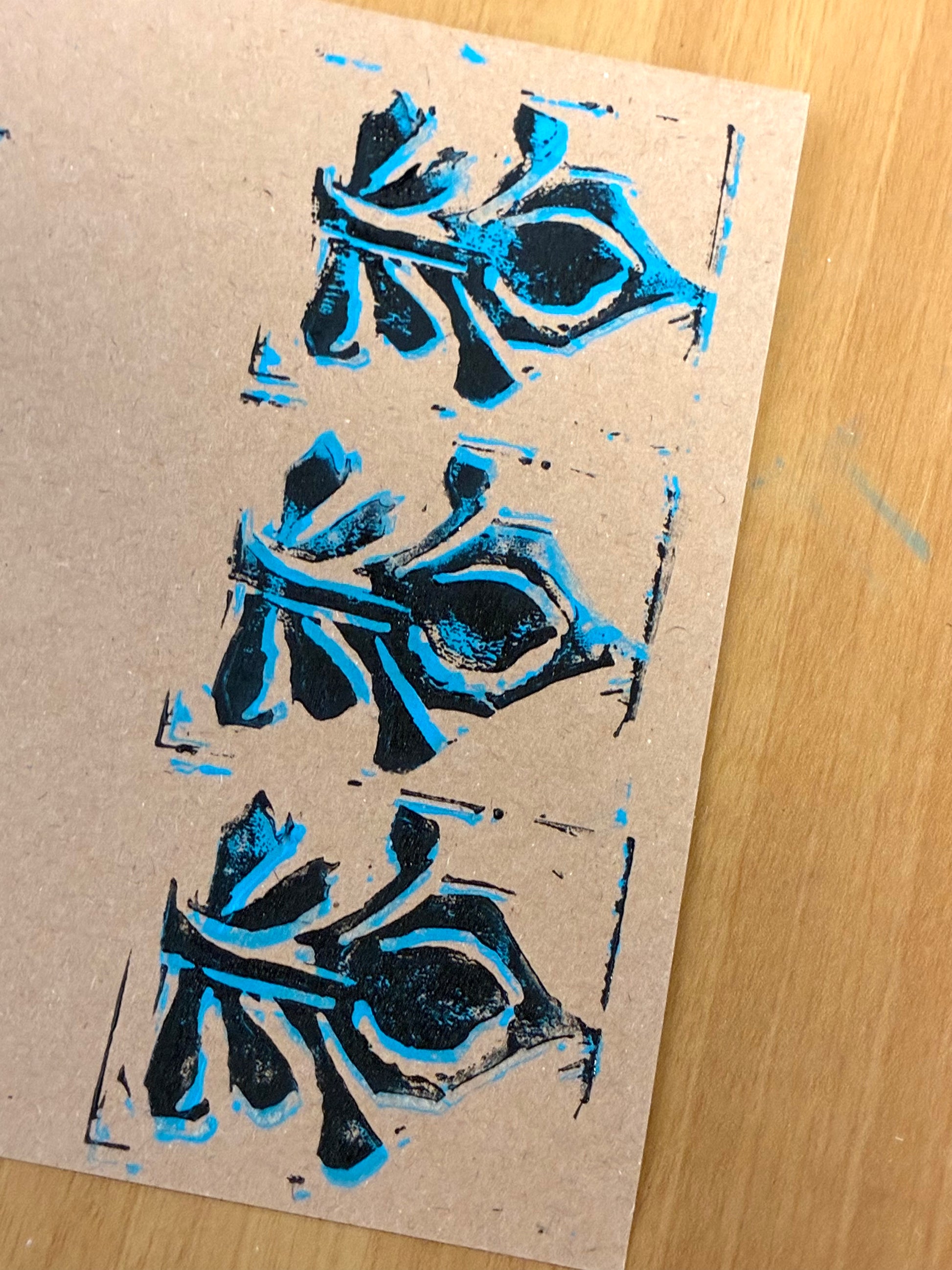 A close-up of a handmade art print featuring three stylized black ink calligraphic characters outlined with bright blue ink, printed on a textured brown kraft paper sheet over a light wood background.