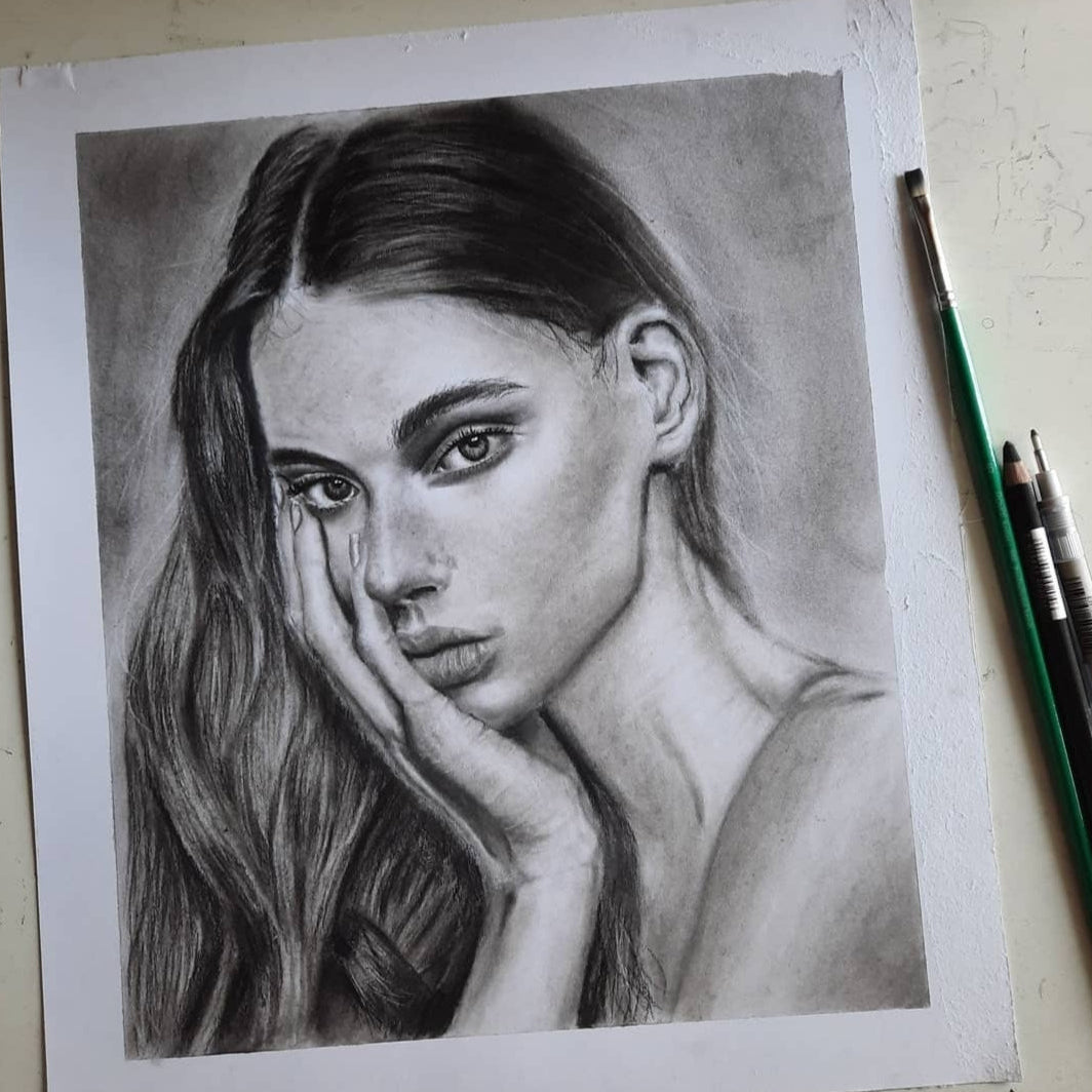 A detailed black and white pencil sketch of a woman's portrait on textured paper, with three drawing pencils placed on the right side of the paper.